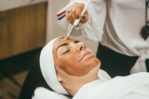 一名女性躺着接受美容护肤面膜疗程，美容师正用刷子将棕色面膜涂抹于其面部